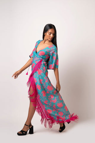 Walking pose of model in asymmetric blue floral wrap dress, one leg forward, showing fringe and flare sleeves in motion.